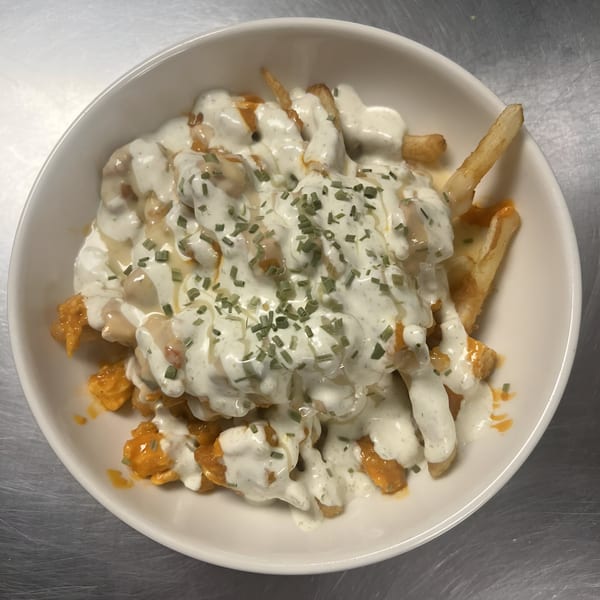 A white bowl filled with golden fries, topped with creamy white sauce, orange buffalo sauce, and chives.