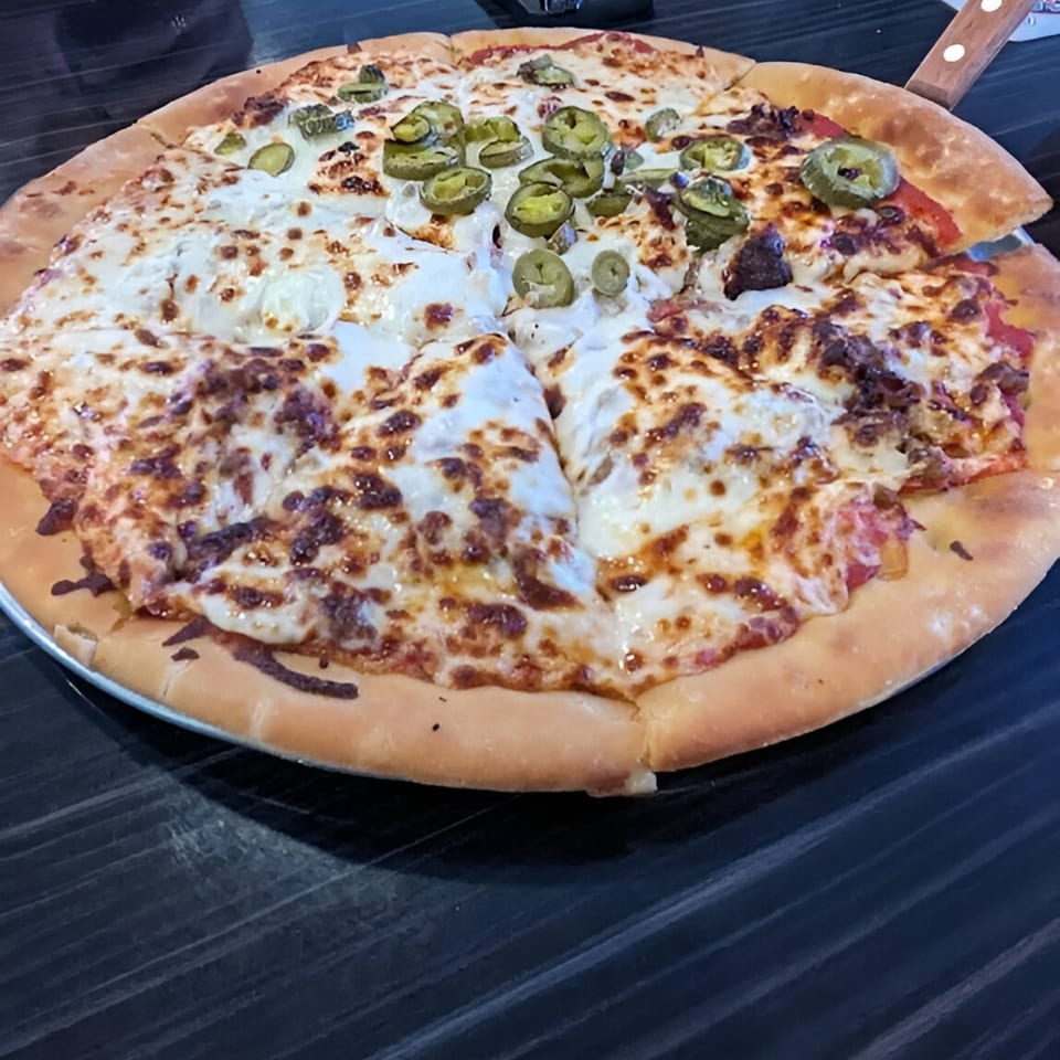 A large, round pizza with melted cheese, jalapeños, and meat, on a dark table.