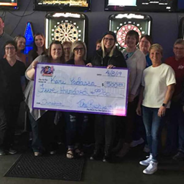A group of people holding a large check for $500, with dartboards in the background at The Rush Bar & Grill.
