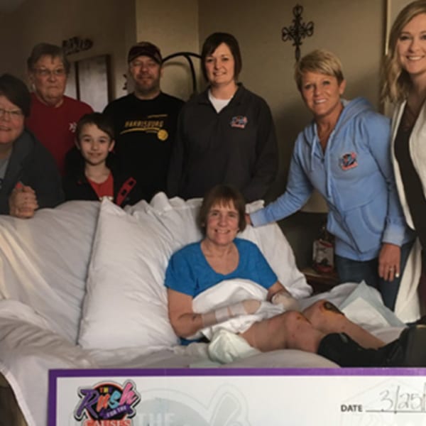 A group of people gathered around a woman in bed, holding a large check from The Rush Bar & Grill.