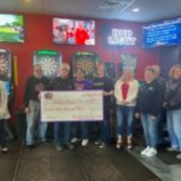 Group of people holding a large check inside The Rush Bar & Grill, with dartboards and TVs in the background.