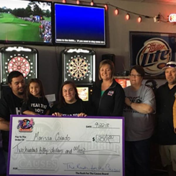 Six people, including a child, hold a large check for $250 in a bar with dartboards and TVs.