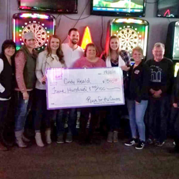 A group of people holding a large check for "Rush for the Causes" in a bar with dartboards.