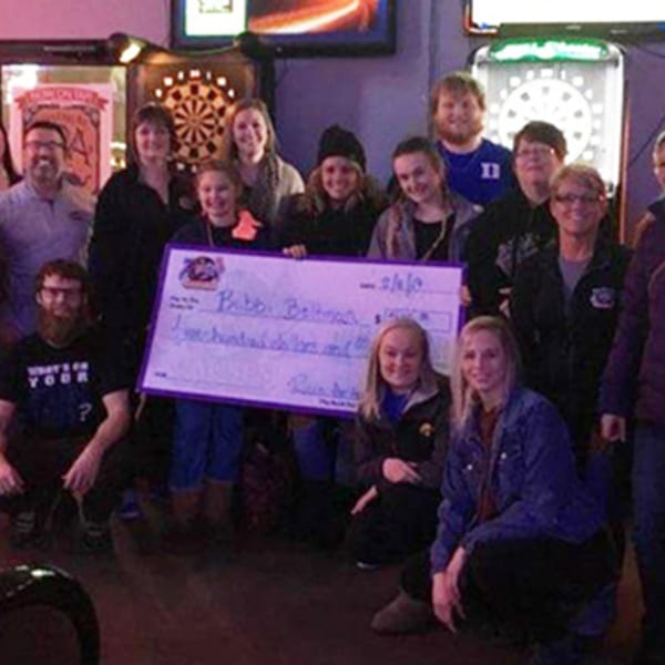 A group of people, some kneeling, holding a large check in a bar with dartboards.
