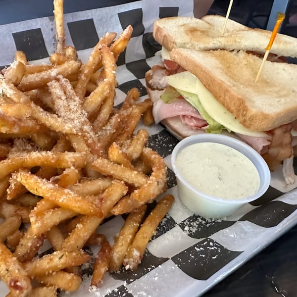 A club sandwich with fries and a side of dipping sauce on checkered paper at The Rush Bar & Grill.
