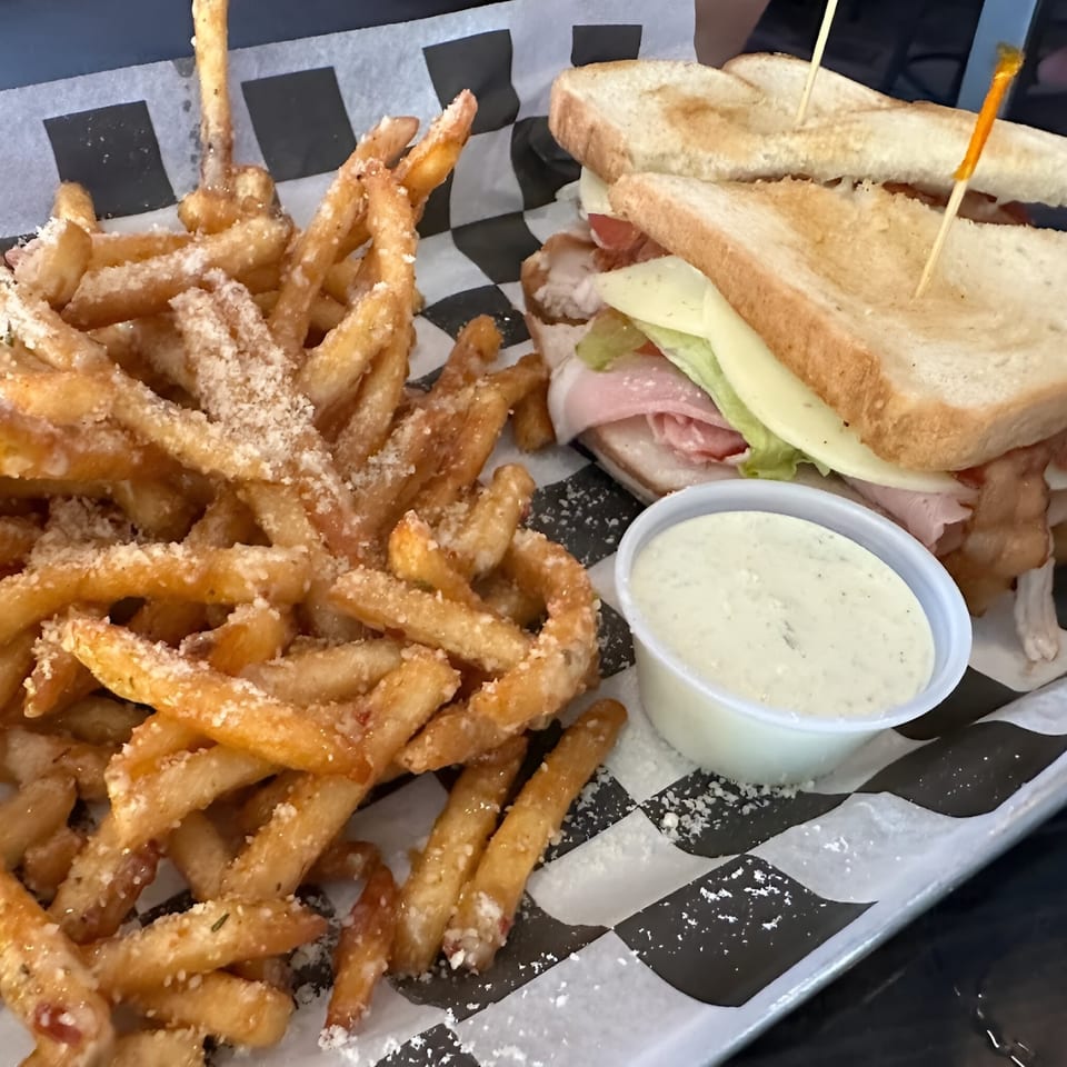 A club sandwich with fries and a side of dipping sauce on checkered paper at The Rush Bar & Grill.