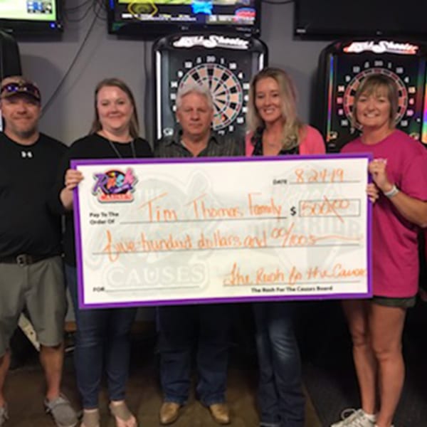 Five people holding a large check for $500 to the Tim Thomas family, with dartboards in the background.