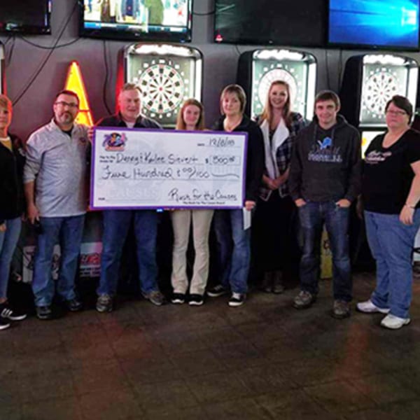 A group of people holding a large check for "Rush for the Cures" in a bar with dartboards.