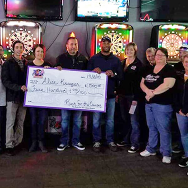 A group of people holding a large check for $500, standing in front of dartboards and TVs.