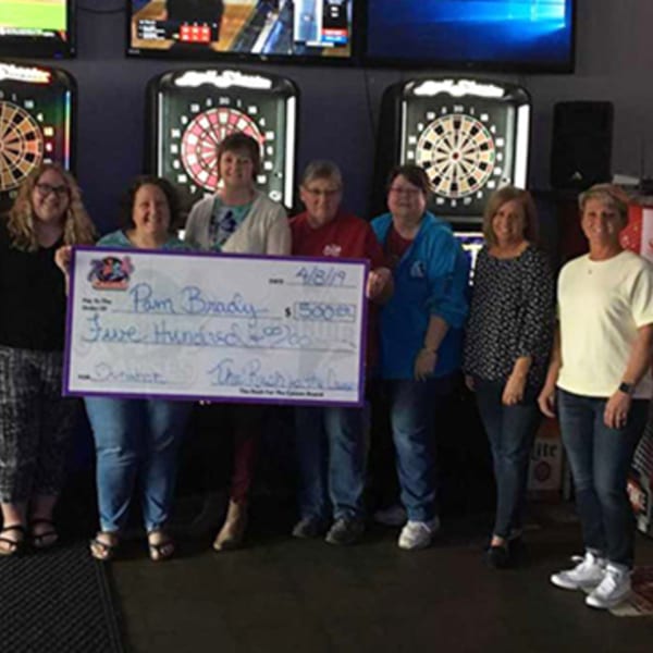 Seven women holding a large check for $500, standing in front of dartboards at The Rush Bar & Grill.