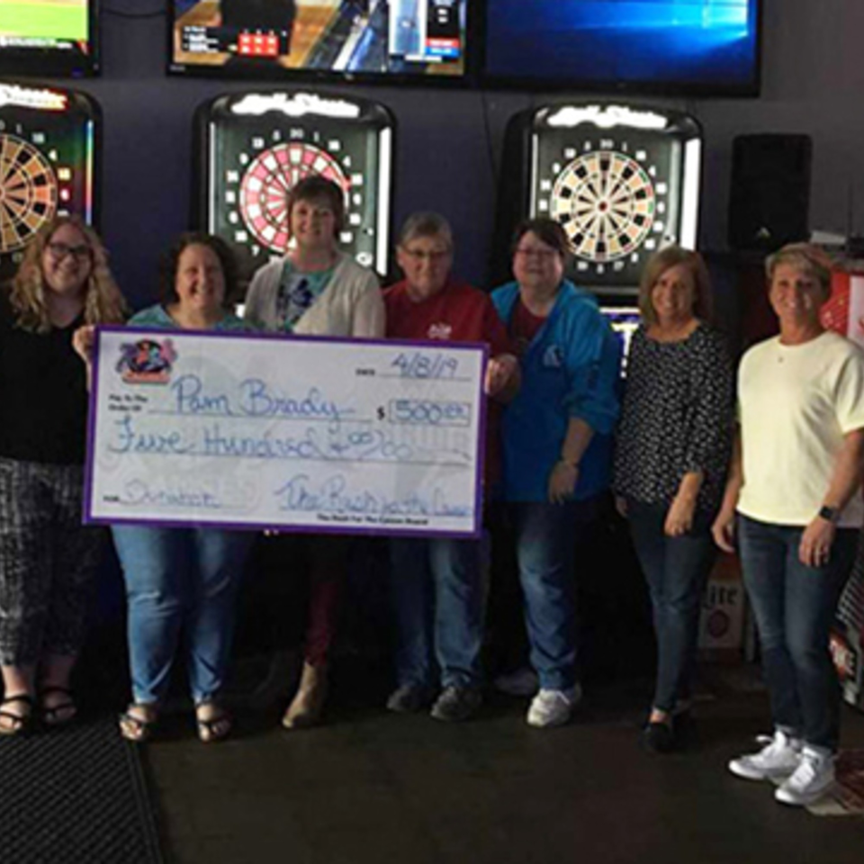 Seven women holding a large check for $500, standing in front of dartboards at The Rush Bar & Grill.