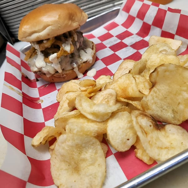 A burger with onion rings and white sauce, served with potato chips on red and white checkered paper.